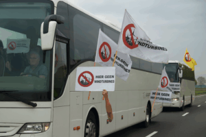 Twee witte bussen vol met protesterende dorpsbewoners onderweg naar Den Haag. Uit de ramen steken borden en vlaggen met het logo en de tekst “Hier geen windturbines”. De sfeer is vastberaden en actiebereid.
